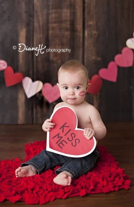 baby sitting on heart shaped rug on the floor holding a kiss me sign - Baby's First Valentine's Day Photo shoot