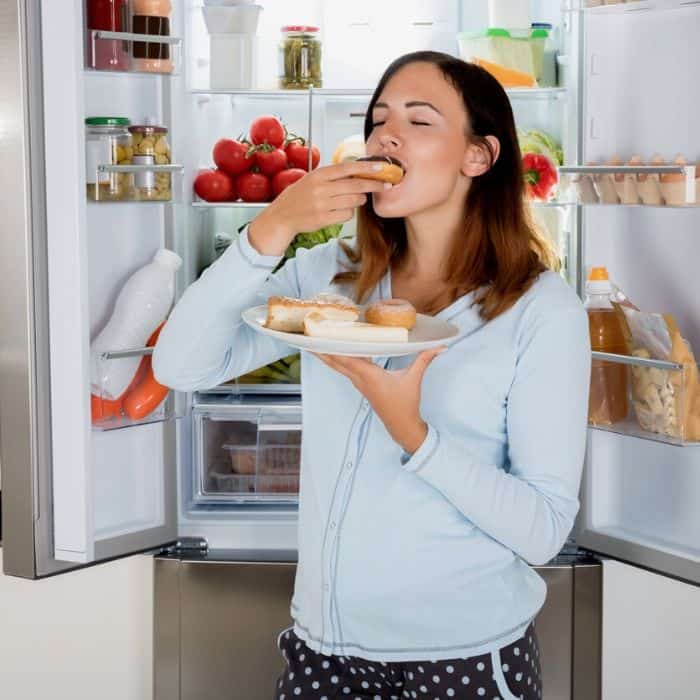 newly pregnant woman eating food in front of open fridge