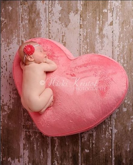 Newborn sleeping on pink fuzzy heart pillow with flower headband on