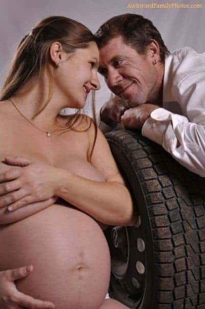 Bare skinned pregnant woman covers breasts with arm while gazing into eyes of man leaning on a tire.