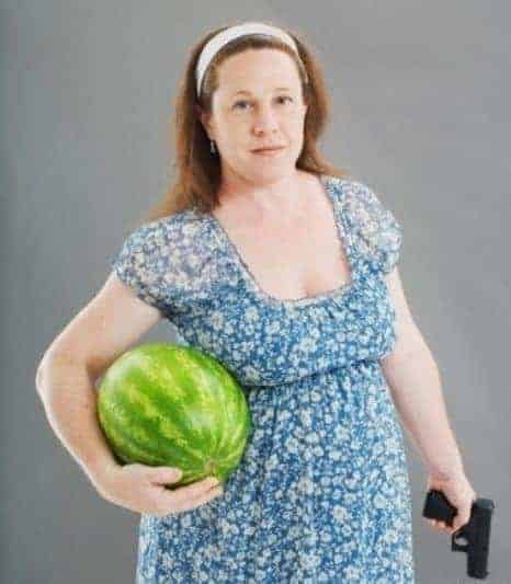 Woman dressed in blue flowery dress smiles at camera while holding a watermelon in one hand and a gun in the other.