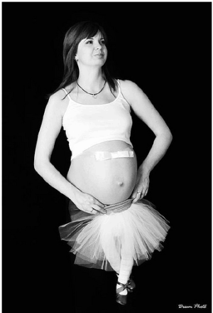 Black and white image of woman with shirt pulled up over bare bump. Small bow is on her bump, and she's holding a child-sized tutu with stuffed tights for legs wearing ballet shoes up below bump giving the illusion she has very tiny legs.