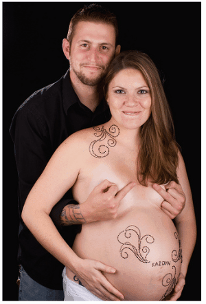 Couple stands facing camera, him behind her covering her bare breasts, flipping the bird with both hands, she topless with body paint. Both smiling mischievously.