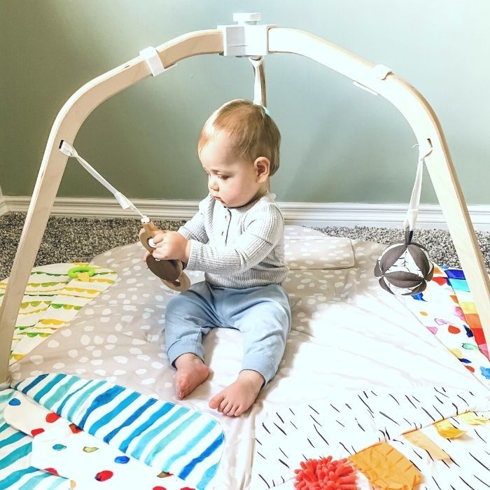 Baby sits on Lovevery play mat while holding attached wooden batting ring.