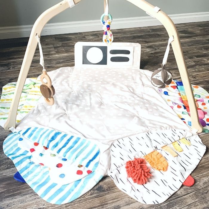 Photo of Lovevery play gym set up and unfolded on the floor. The 5 zones of play are open exposing different flaps, colors, textures, and cards. The wooden batting right, organic teether, and Montissori ball all hang from the wooden legs.