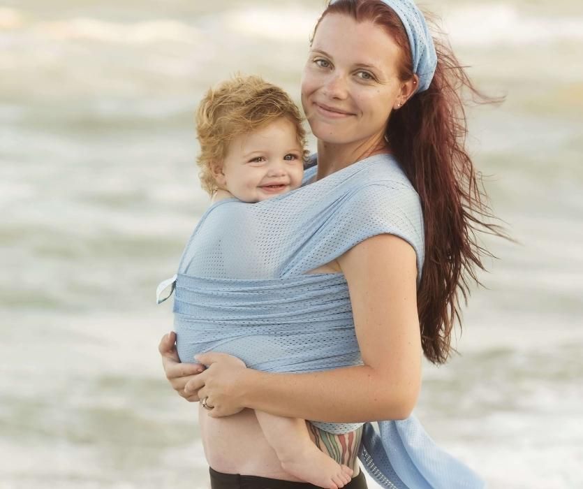 Người mẹ trên bãi biển mỉm cười trước máy ảnh mặc cho đứa trẻ hạnh phúc trong Chiếc khăn quấn em bé bên bờ biển