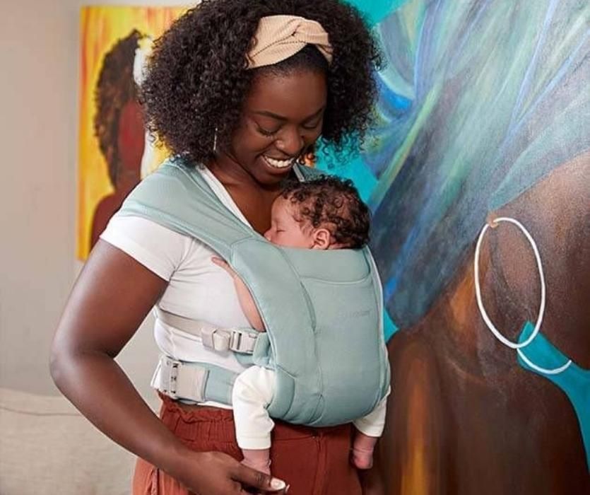 Mom standing in front of colorful wall smiling down at an infant in Ergobaby Embrace Soft Air Mesh carrier.