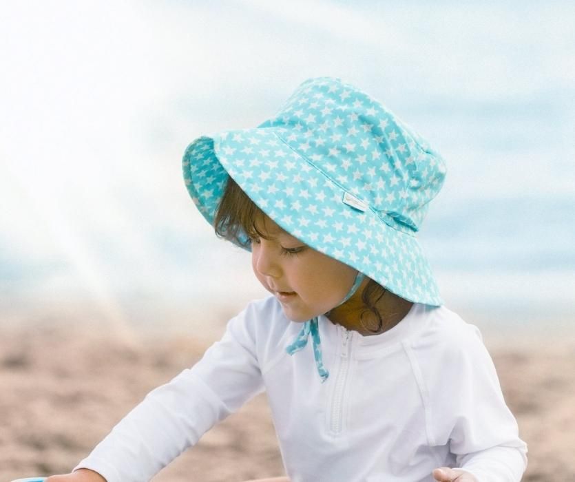 Bé mặc áo chống nắng màu trắng và đội mũ che nắng màu xanh với những ngôi sao trắng đang chơi đùa trên cát.