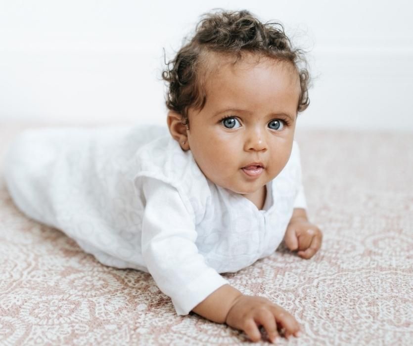 Baby laying on their stomach on neutral carpet wearing white and gray Halo Cotton Muslin Sleep Sack