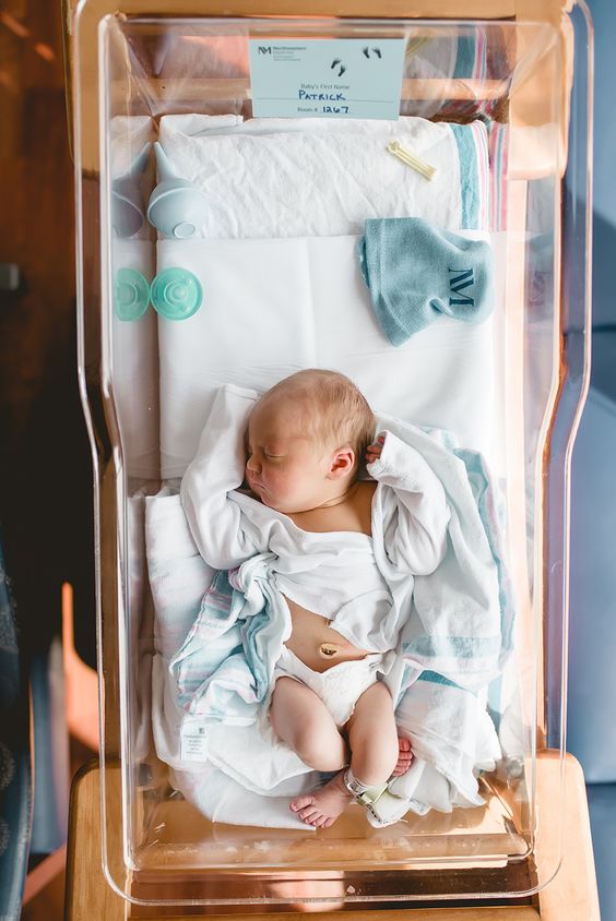 Color photo from above of un-swaddled newborn sleeping in hospital bassinet with legs tucked up and arms above their head.