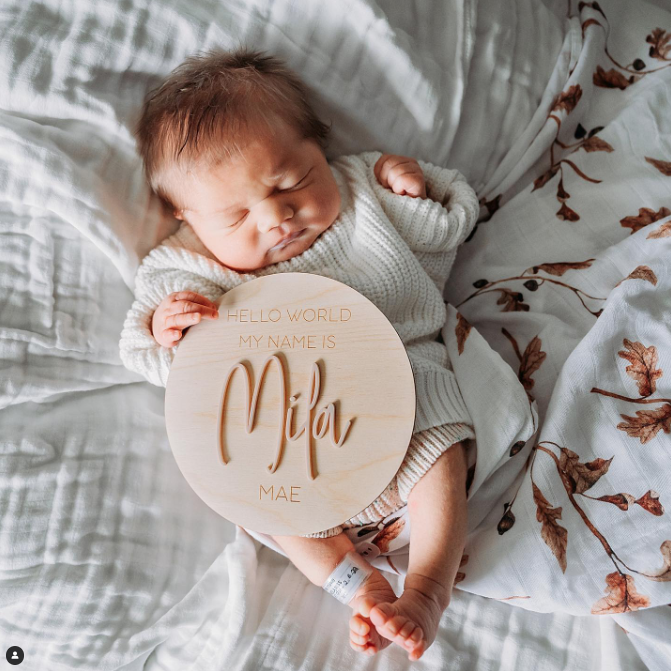 Color photo from above of sleeping newborn holding a sign that reads: Hello world, my name is Mila Mae. 