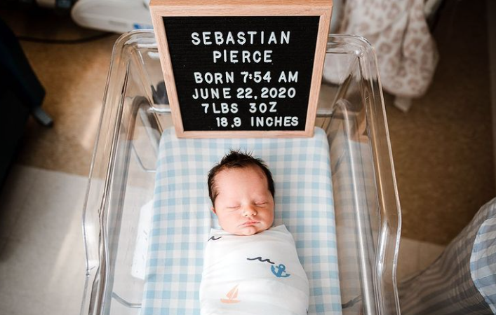 Color photo taken from above showing sleeping newborn with letter board in his hospital bassinet with name and birth details listed.