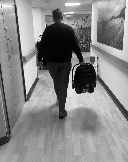 Black and white photo of parent carrying infant in bucket carseat through hospital halls.