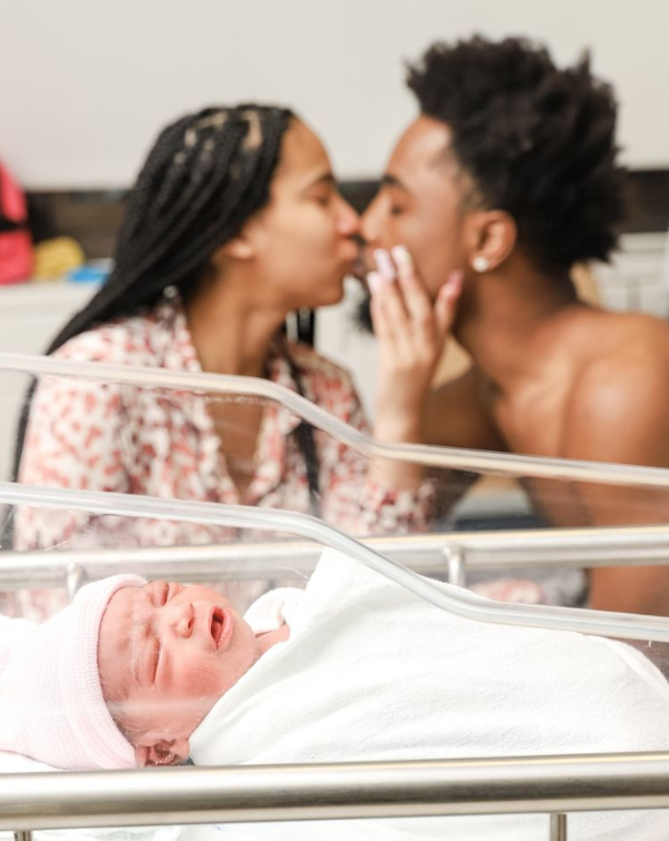 Color photo of mom and dad kissing in the background of a photo with a sleeping swaddled baby in the foreground. 