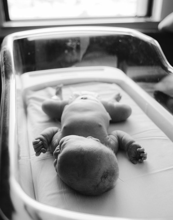 Black and white photo of baby sleeping in hospital bassinet wearing nothing but a diaper.