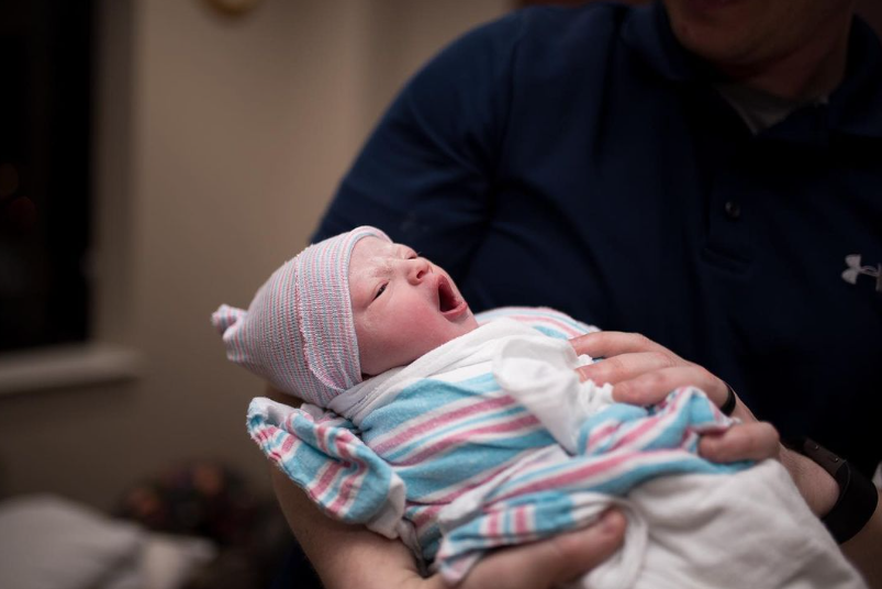 Color photo of swaddled newborn yawning while being held by someone in a dark blue shirt. 