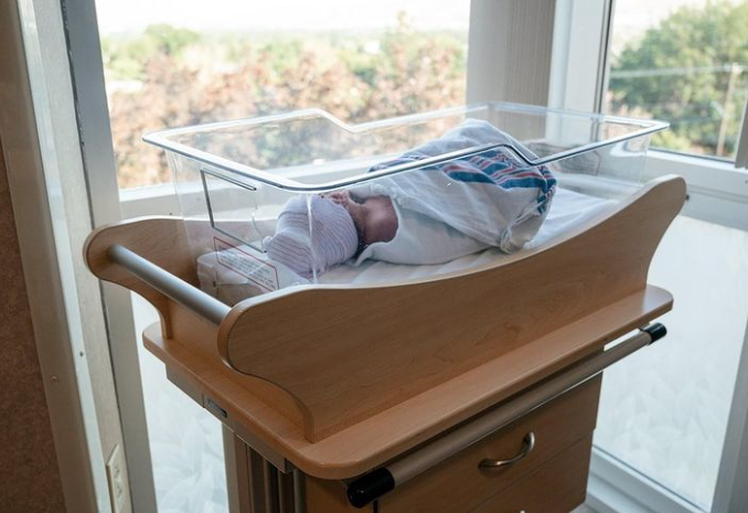 Color photo of swaddled baby sleeping in hospital bassinet in front of two windows.