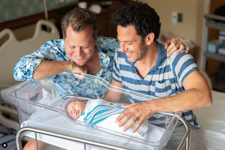 Color photo of two dads dressed in blue smiling down at their newborn swaddled in a hospital bassinet.