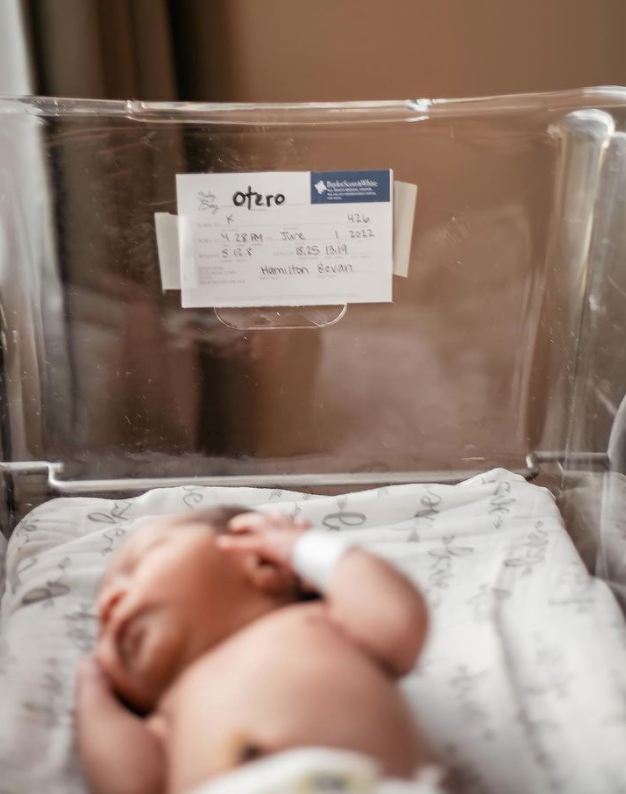 Color photo of sign in the hospital bassinet with birth details while newborn sleeps in foreground.