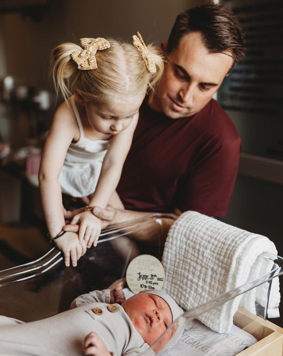 Color photo of older sister leaning over hospital bassinet with help of dad looking down at her new sibling.