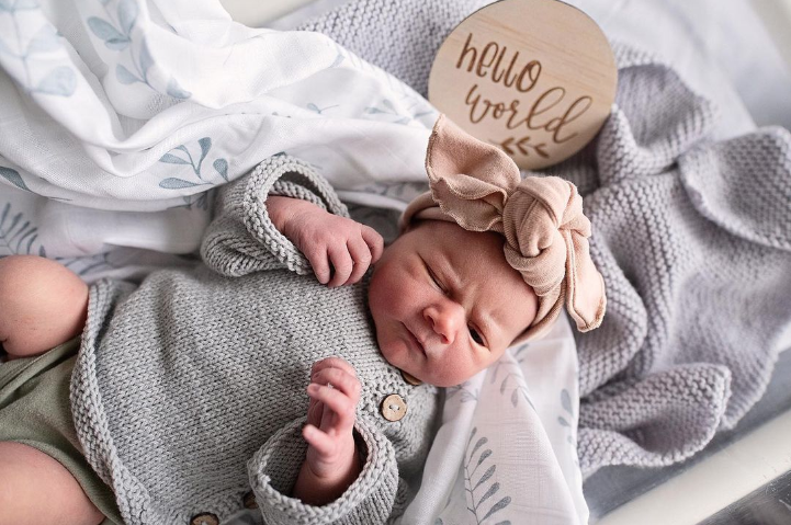 Color photo from above of newborn laying on blankets with pink bow and sign that reads: hello world. 