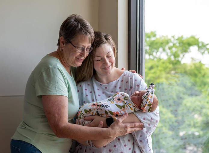 Color photo of grandma, mom, and new baby standing in front of a window, smiling down at the baby.