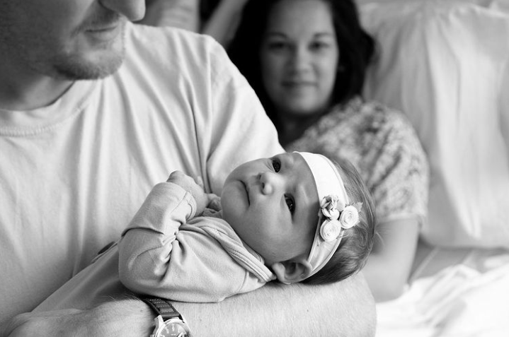 Black and white photo of dad holding newborn wearing a flower headband while mom smiles in the background.