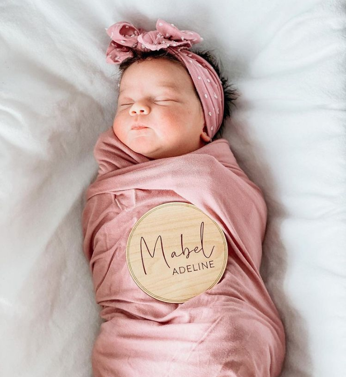 Color photo from above showing sleeping baby swaddled in pink, wearing a pink and white polk-a-dot headband with a sign on her chest that says, "Mabel Adeline"