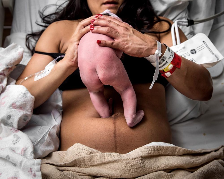 Color photo of mom holding naked newborn standing on her belly.