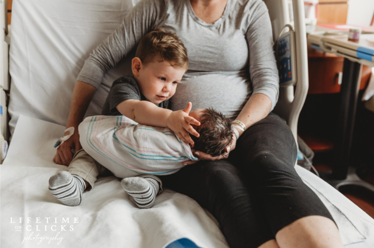 Color photo of toddler brother reaching for newborn sibling's head while mom helps hold everyone on the hospital bed.