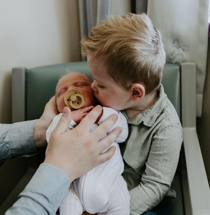 Color phot showing older brother kissing head of newborn sibling while parent holds newborn baby in his lap. 