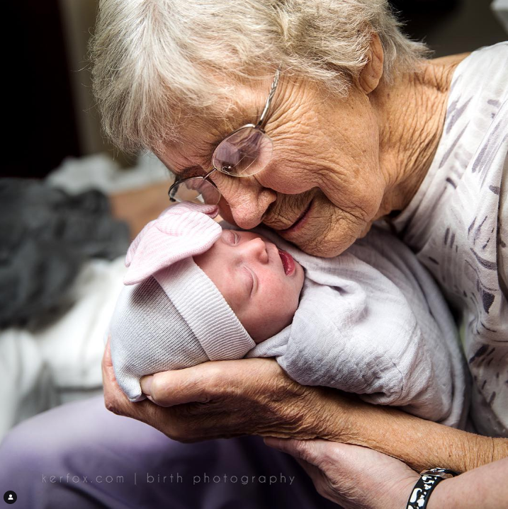 Color photo of elderly grandparent holding infant cheek to cheek.