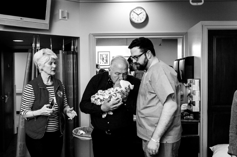 Black and white photo of grandparents meeting new baby in the hospital while dad looks on.