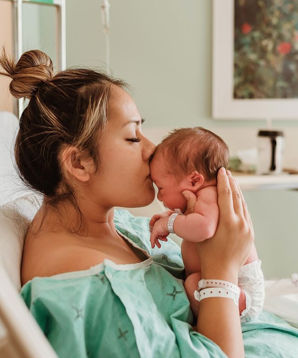 Profile color photo of mom holding up newborn wearing nothing but a diaper getting kissed on the head.