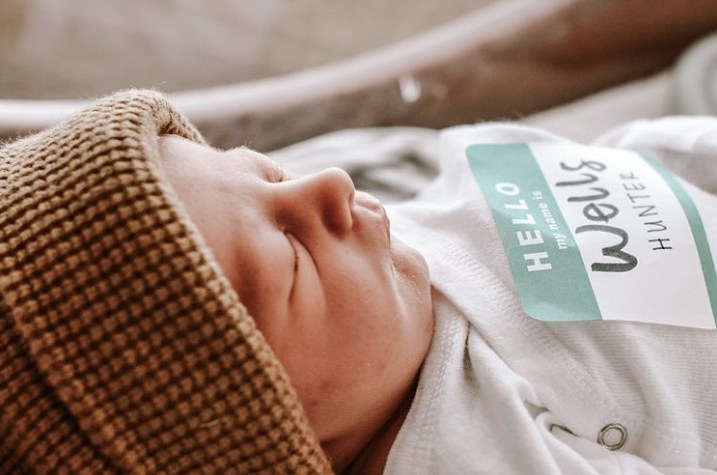 Close up color photo of newborn sleeping with sticker on their shirt that says, "Hello, my name is Wells Hunter"