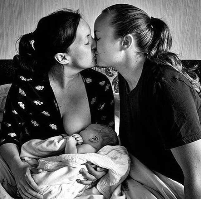 Black and white image of moms kissing over sleeping baby being nursed.