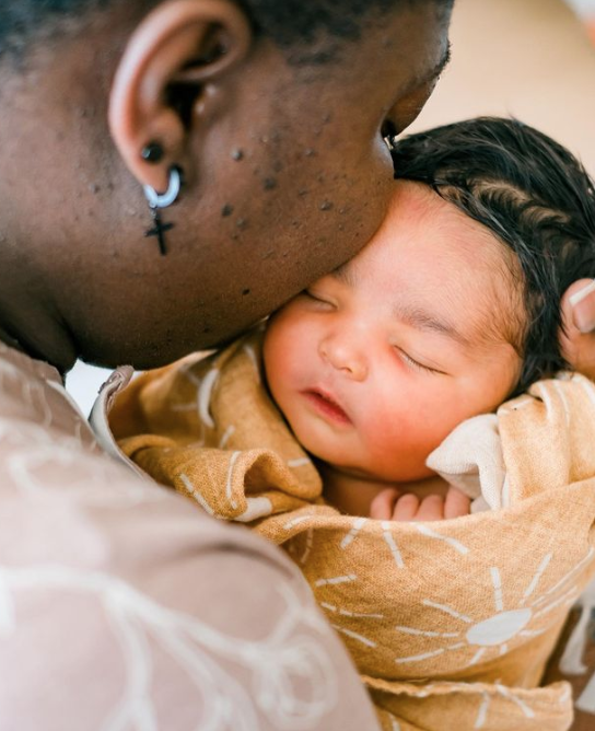 Color photo of mother holding her cheek to the forehead of her newborn swaddled in yellow swaddle with white suns.