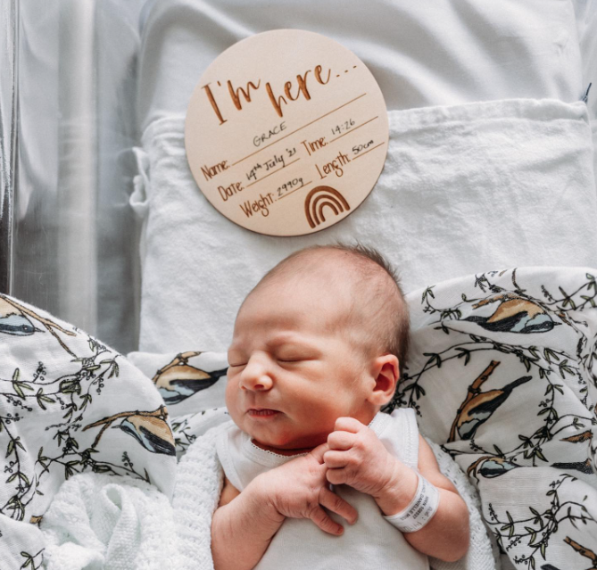 Color photo from above showing half a sleeping newborn and a sign that says, "I'm here..." with name, and birth details filled in.