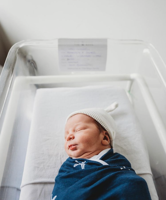 Color photo of baby sleeping in hospital bassinet wearing dark blue swaddle and white hat. 