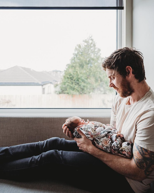 Color photo of dad holding newborn swaddled in flower print sitting on a bench in front of a window.