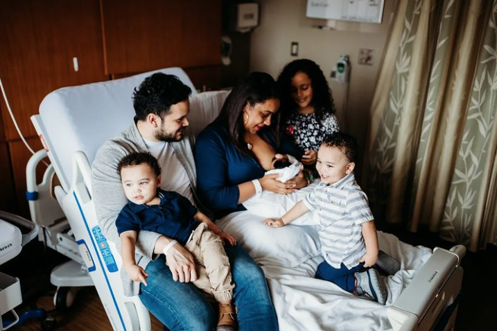 Color photo of three older kids sitting on hospital bed with mom and dad while mom nurses the newborn.