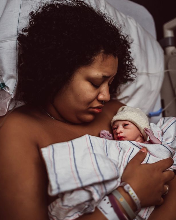 Color photo of mother looking down at newborn swaddled in hospital blanket wearing a hat.