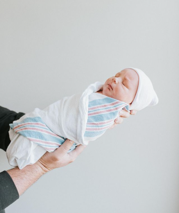 Color photo of partner holding baby up in front of white wall while they sleep, swaddled and in a hat. 