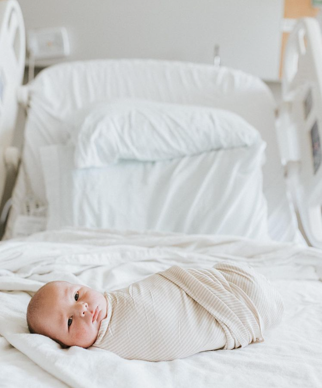 Color photo of newborn laying on white hospital bed swaddled in light colored swaddle looking at camera.