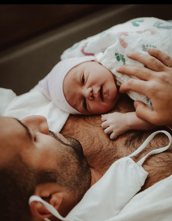 Color photo from above of dad lying down doing skin to skin while baby looks at camera.