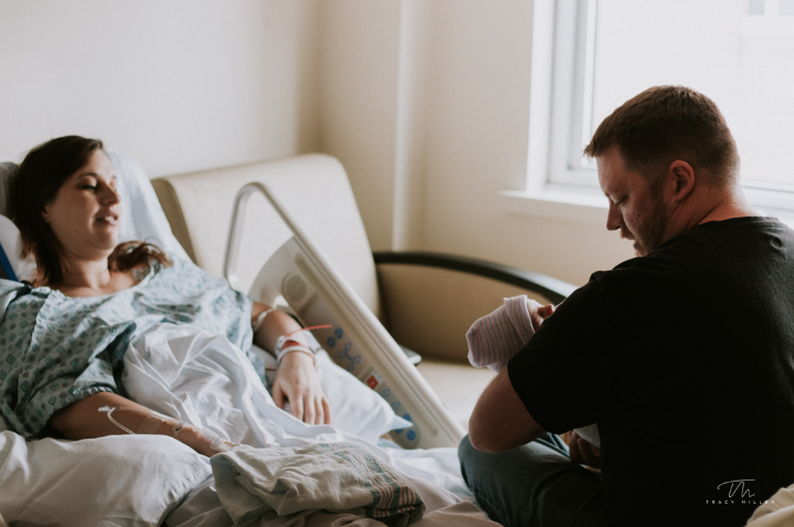 Color photo of dad admiring the baby he's holding while mom lays in hospital bed looking on.