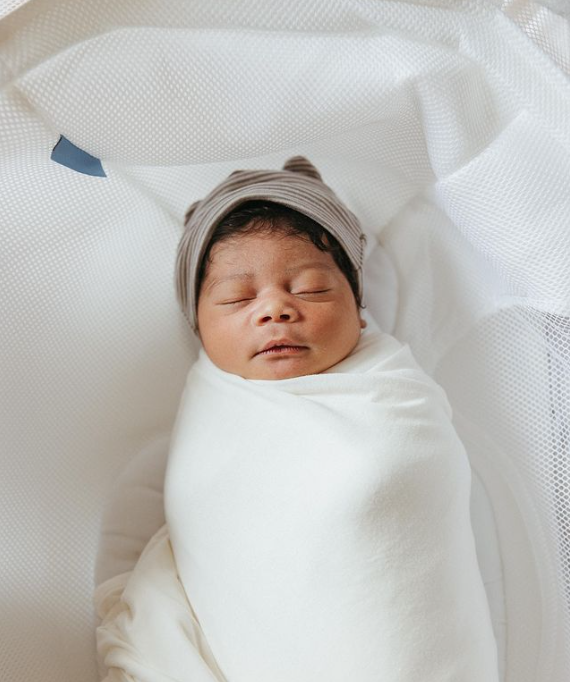 Color photo of baby sleeping in a white swaddle, wearing a bear hat.