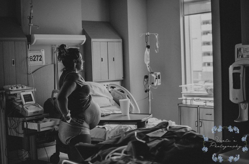 Black and white photo of pregnant mom in hospital room looking out window while she labors