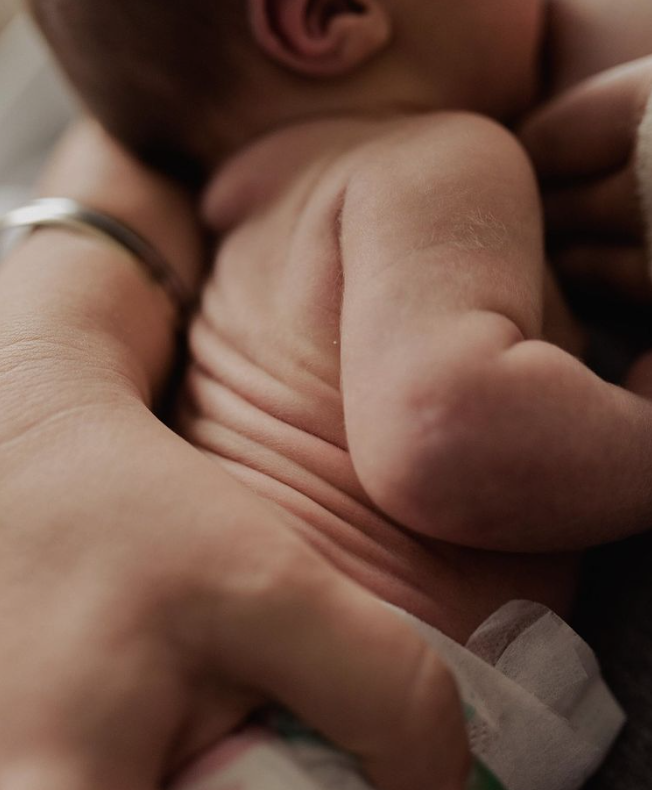 Color photo of close up of mom's hand and baby's wrinkled back, and peach fuzz shoulders.