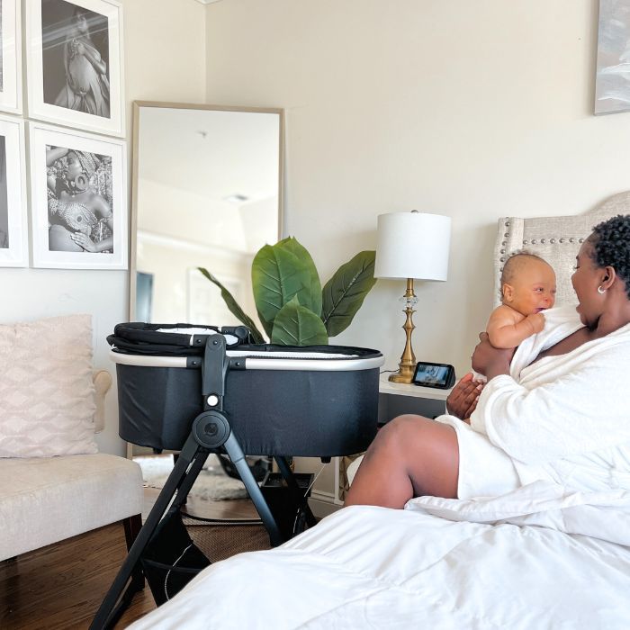 Woman sitting on bed with Mockingbird bassinet with stand next to her.
